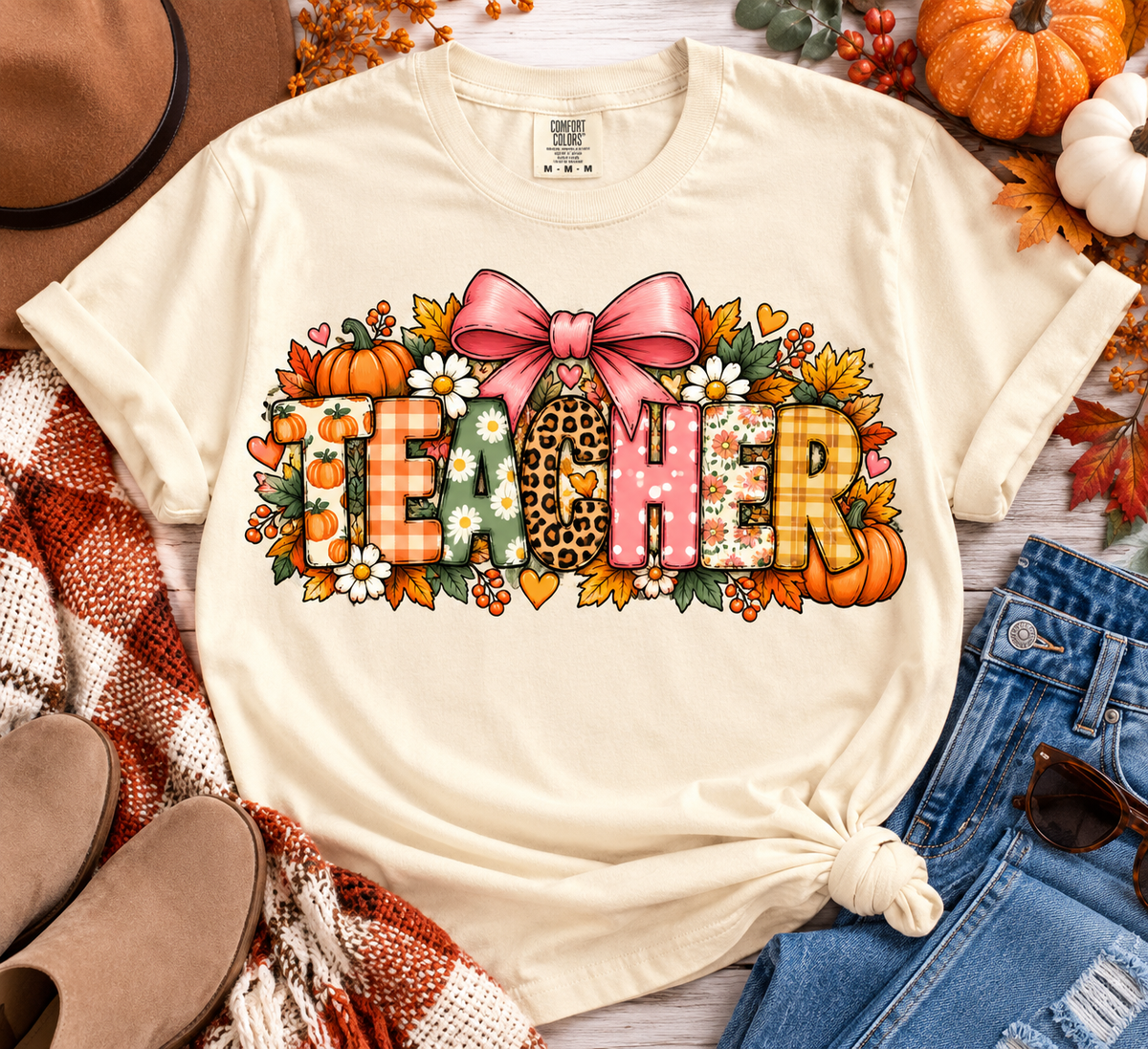 Ivory Comfort Colors fall teacher shirt with patterned TEACHER lettering, pink bow, pumpkins, florals and autumn details styled in a cozy flat lay.