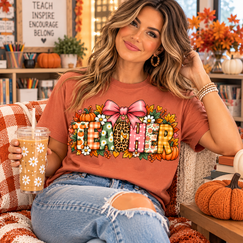 Teacher wearing a Terracotta Comfort Colors fall TEACHER shirt with colorful patterned lettering and pink bow in an autumn-decorated classroom.
