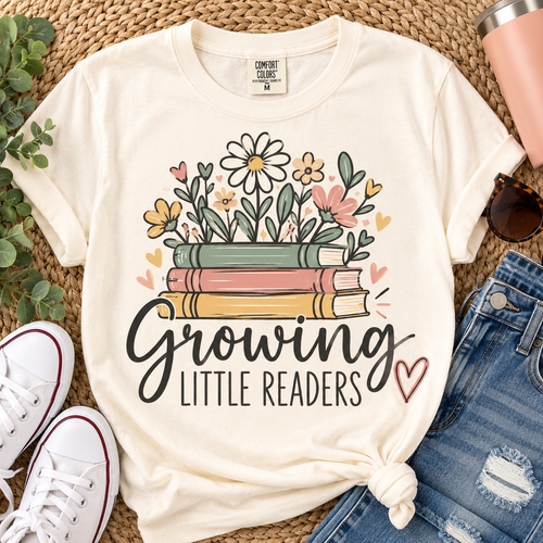 Ivory Comfort Colors Growing Little Readers teacher shirt with colorful books and flowers styled in a flat lay with jeans and sneakers.