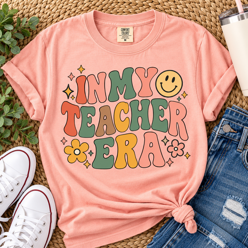 Blossom Comfort Colors In My Teacher Era shirt with retro lettering, smiley face and floral details styled in a teacher-inspired flat lay.