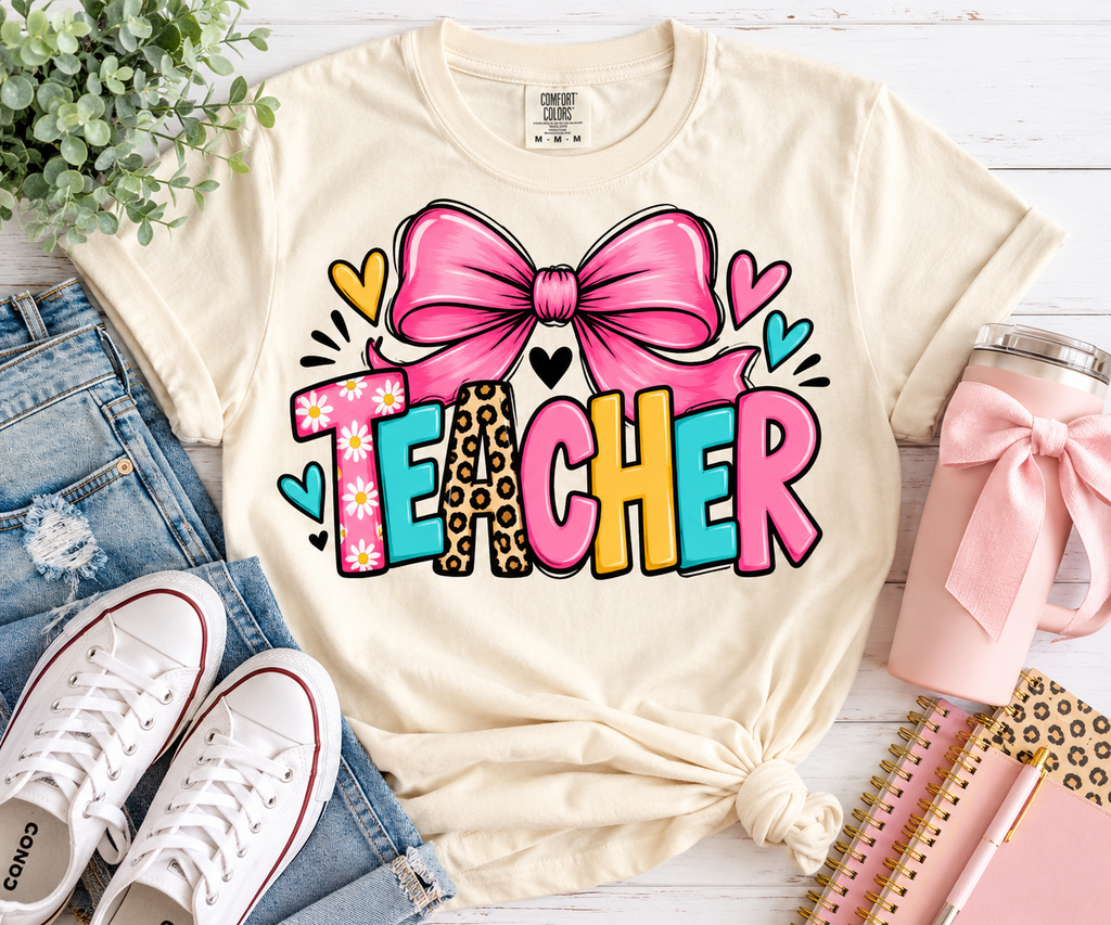 Ivory Comfort Colors teacher shirt with colorful TEACHER lettering and a pink bow styled with jeans, sneakers, and teacher accessories.