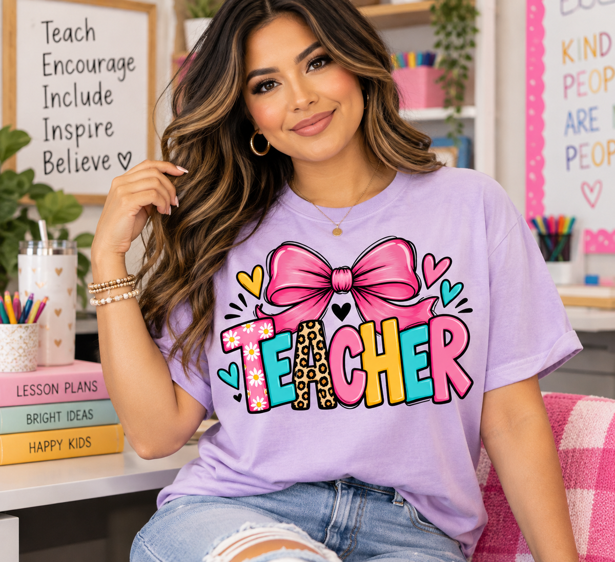 Woman wearing an Orchid Comfort Colors teacher shirt featuring colorful TEACHER lettering and a large pink bow in a classroom setting.