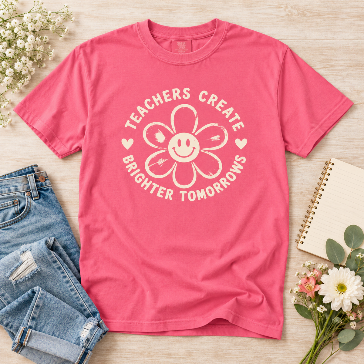 Crunchberry pink teacher T-shirt laid flat with a cream smiling flower graphic and “Teachers Create Brighter Tomorrows” printed on the front.
